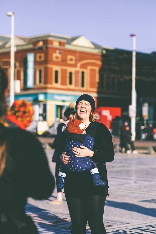 mother laughing while babywearing in a connecta baby carrier. Baby is wearing a fox hat and is snuggled into mum. they are outside on a bright autumn day