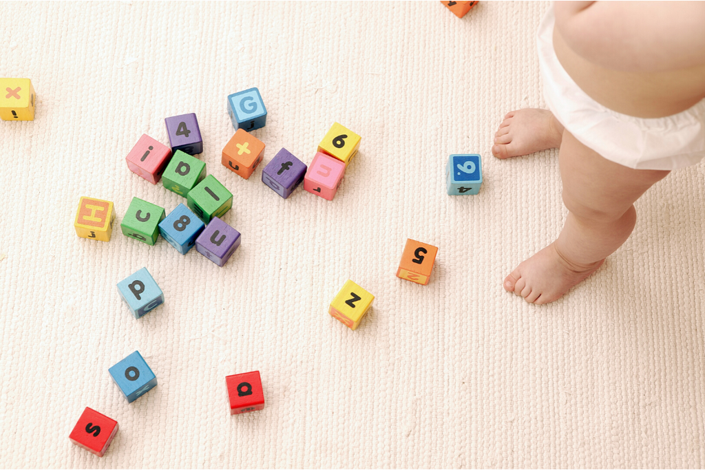 toddler standing in their nappy looking down over a range of coloured letter blocks scattered on the floor.